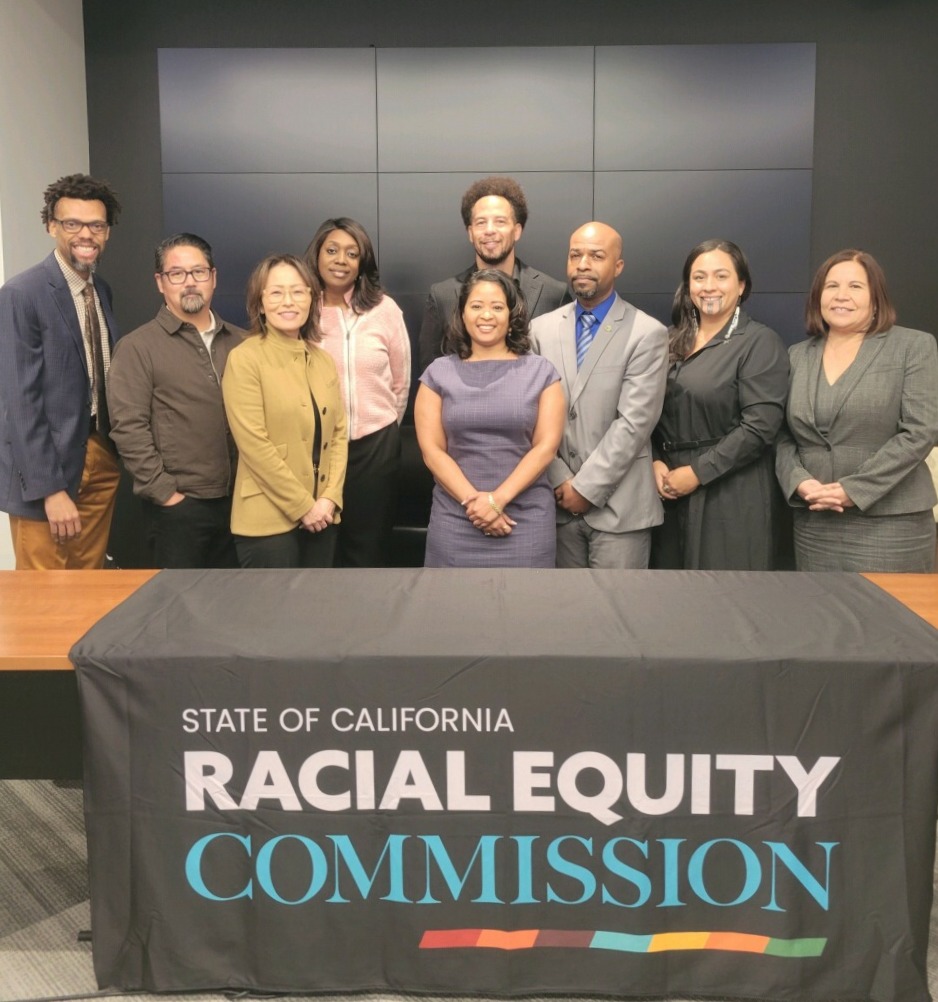 Commissioners pose behind table draped with black California State Racial Equity Commission table cloth / banner.