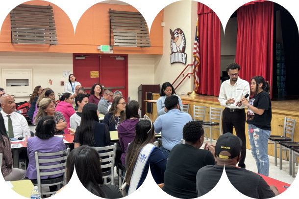 A community gathering in a school auditorium with red curtains and an American flag on stage. Attendees sit at tables while two presenters stand at the front, speaking to the group. The atmosphere appears collaborative and interactive.